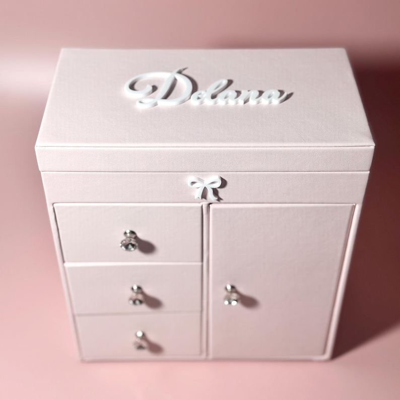 May include: A light pink jewelry box featuring a white script name Delana on the top lid, a small white bow accent on the front, three drawers with clear crystal-like knobs, and a side cabinet door with a matching crystal-like knob.