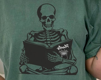 Puede incluir: Una camiseta verde con un esqueleto sentado con las piernas cruzadas y leyendo un libro que dice "Plantas".