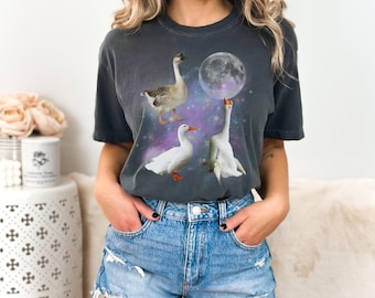 Op de afbeelding: Een donkergrijs T-shirt met een afbeelding van drie ganzen in een galaxie met een maan. De ganzen zijn wit met oranje snavels en poten. De maan is wit met een galaxie achtergrond.