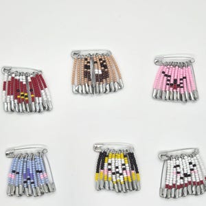 May include: A collection of six beaded safety pin brooches. Each brooch features a unique design with colorful beads, including red, white, pink, black, yellow, and blue. The brooches are arranged on a white surface.