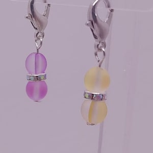 Pode incluir: Dois pingentes de charme com fechos prateados. Os pingentes são feitos de contas de vidro fosco com uma faixa prateada no meio. Um pingente é roxo claro e o outro é amarelo claro.