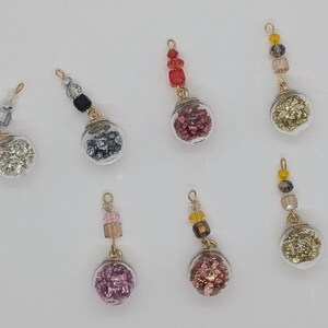 Puede incluir: Siete adornos de vidrio con alambre dorado y cuentas que cuelgan de ellos. Los adornos están llenos de purpurina de diferentes colores. Los colores incluyen plata, negro, rojo, oro, rosa y marrón.