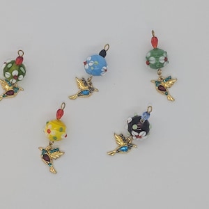 Puede incluir: Cinco abalorios de tono dorado con cuentas de vidrio de colores y detalles de colibrí. Cada abalorio presenta un patrón floral diferente en la cuenta.