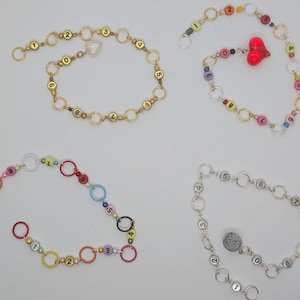 以下が含まれることがあります： 番号の付いたチャームが付いた、カラフルなビーズネックレスが4本。各ネックレスは異なるカラーパターンで、チャームが端に付いています。チャームは、小さなハート、小さな鳥、小さな花、小さな石です。