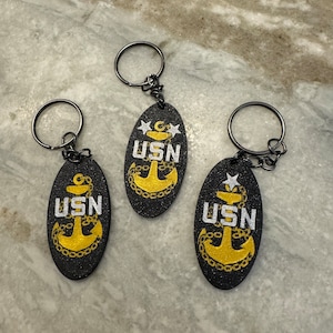 May include: Three black keychains with silver key rings. Each keychain features a black oval charm with a gold anchor and the text "USN" in white lettering.