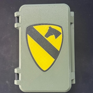 May include: An olive green rectangular case with a hinged lid. The case features a yellow shield with a black stripe and a black horse head silhouette. The case is closed and appears to be made of plastic.