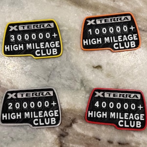 May include: Four rectangular patches with the text "XTERRA HIGH MILEAGE CLUB" and mileage numbers: 100000+, 200000+, 300000+, and 400000+. Each patch has a different colored border: orange, yellow, gray, and red.