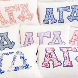 Puede incluir: Cojines rectangulares blancos con letras griegas bordadas en varios patrones florales y colores, incluyendo rosa, azul y morado. Las letras están delineadas en colores coordinados, creando un diseño decorativo y personalizado.
