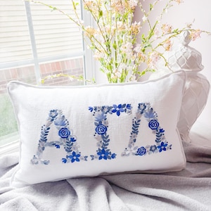 Op de afbeelding: Een wit rechthoekig kussen met de geborduurde letters "ATA" in blauwe en grijze bloemmotieven. Het kussen ligt op een grijze deken bij een raam met een vaas met bloemen.