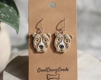 Puede incluir: Un par de pendientes colgantes con un rostro de perro pit bull marrón y blanco en cada pendiente. Los pendientes están en una tarjeta marrón con el texto "Good Dawg Goods" y un gráfico de hueso.