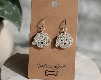 Può includere: Un paio di orecchini pendenti con una testa di cane in ceramica bianca con occhi e naso neri. Gli orecchini sono esposti su una carta marrone con il testo "Good Dawg Goods" e un grafico di un osso.