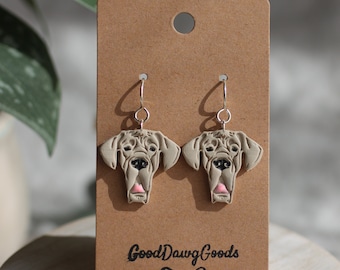 Puede incluir: Un par de pendientes de plata con un dije de cabeza de perro Gran Danés marrón claro. Los pendientes se muestran en una tarjeta marrón con el texto "Good Dawg Goods" y un icono de hueso.