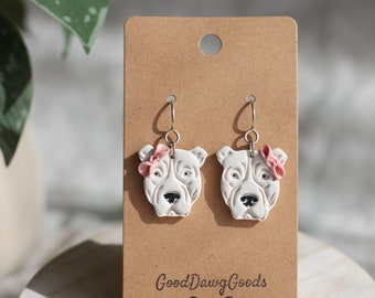 Puede incluir: Un par de pendientes colgantes de perro blanco con lazos rosas en una tarjeta marrón con el texto "GoodDawgGoods" y un icono de hueso.