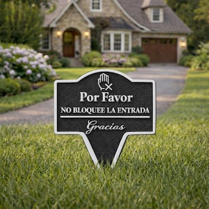 May include: A black and white metal lawn sign with the words "Por Favor, NO BLOQUEE LA ENTRADA, Gracias". The sign has a hand symbol with an X through it. The sign is in a grassy yard in front of a house.