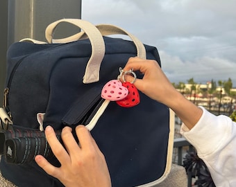 May include: A blue canvas duffel bag with a white zipper and two red and pink heart-shaped keychains attached to the zipper pull.