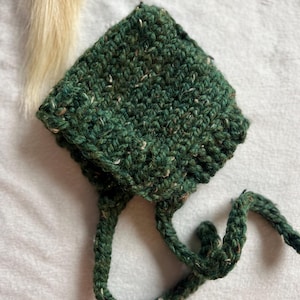 May include: A handmade, green crochet bonnet with speckled white and brown yarn. The bonnet has a square crown and long, braided ties. The textured knitwear is displayed on a soft, white surface.