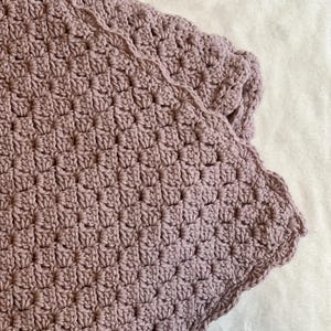 Può includere: Primo piano di una coperta all'uncinetto color rosa antico. La coperta ha un motivo strutturato e un bordo smerlato. Il filato appare morbido e le cuciture sono fitte.