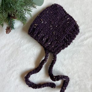 May include: A knitted purple baby bonnet with speckled yarn. The bonnet has a rounded crown and long, braided ties. The yarn is a deep purple with flecks of cream and tan. The bonnet is likely handmade and suitable for a newborn.