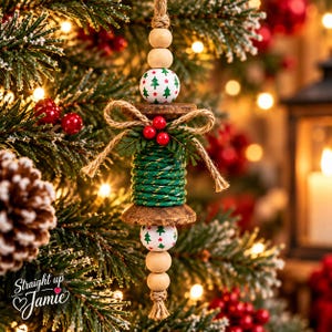 May include: A hanging Christmas ornament featuring a central wooden spool wrapped in green and gold twine, accented with a jute bow, three red berries, and green pine needles. The ornament includes white beads with green trees and red stars.