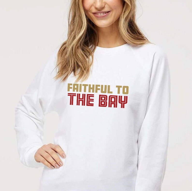 May include: White long-sleeve sweatshirt with the text "Faithful to The Bay" in gold and red lettering.