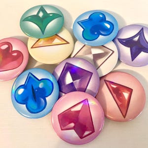 May include: A collection of ten round, colorful buttons with various geometric and abstract designs. The buttons feature a range of colors, including pink, blue, green, and purple, with holographic and iridescent effects. The designs include hearts, diamonds, and abstract shapes.