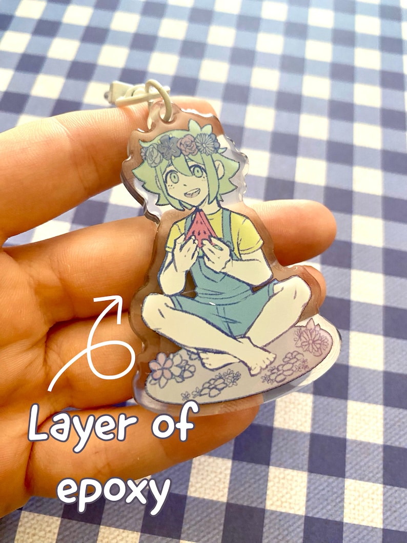 Op de afbeelding: Een transparante acryl sleutelhanger met een cartoonfiguur met groen haar, die een geel shirt en een blauwe overall draagt, een plak watermeloen vasthoudt. Het personage is omringd door bloemen en zit op een wolkachtige vorm. De sleutelhanger is bedekt met een laag epoxy.