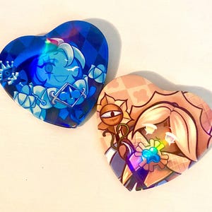 May include: Two heart-shaped pins. One is blue with a diamond pattern and a cartoon character. The other is peach with a floral pattern and a cartoon character. Both pins have a holographic effect.