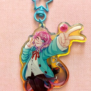 May include: A colorful anime character keychain with a blue star-shaped charm. The character has pink hair, blue eyes, and is wearing a blue jacket with a red bow tie. The character is holding a pink lollipop and making a peace sign with their right hand.