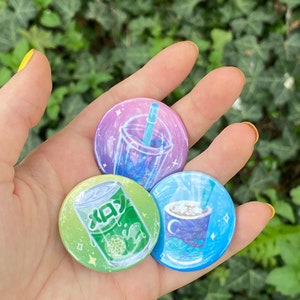 May include: Three round pins with different designs. The first pin is green and yellow with a can of melon soda. The second pin is purple and blue with a cup of boba tea. The third pin is blue with a cup of hot chocolate.