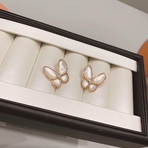 Op de afbeelding: Een paar vlinderoorbellen. De oorbellen hebben een goudkleurig montuur met iriserende, witte vleugels en een kleine, heldere edelsteen in het midden van elke vlinder. Ze worden gepresenteerd in een zwart-witte sieradendoos.