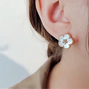 Op de afbeelding: Goudkleurige bloemvormige oorbel met een witte parelmoeren kern en een kralenrand. De oorbel wordt op een oor gedragen en toont het delicate ontwerp en de elegante uitstraling. Het bloemmotief voegt een vleugje verfijning toe.