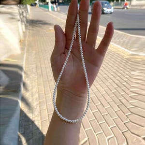 May include: A single strand necklace composed of small, uniform, round white pearls. The necklace is held in a vertical loop, showcasing the consistent size and smooth, reflective surface of each individual pearl bead along the entire length of the strand.