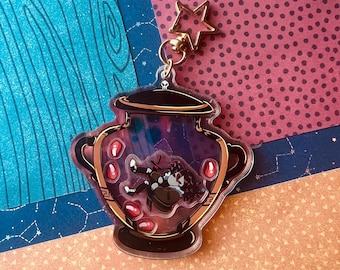 Pode incluir: Um porta-chaves de acrílico transparente em forma de urna decorativa com um fecho dourado em forma de estrela. A urna apresenta uma borda e base castanho-escuras, contendo uma pequena ilustração de uma figura a flutuar entre formas vermelhas.