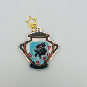 May include: A gold star charm attached to a clear acrylic keychain featuring a cartoon character inside a blue and white urn with red accents.