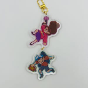 May include: A gold keychain with two clear acrylic charms. The top charm features a cartoon girl with brown hair holding a pink weapon. The bottom charm features a cartoon boy with a blue hat and a red shirt holding a book.