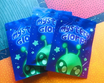 May include: Three rectangular blue foil pouches featuring a green alien graphic and the text "mystery glorps" in a stylized font. The packaging includes small star illustrations scattered around the alien character on a deep blue background.