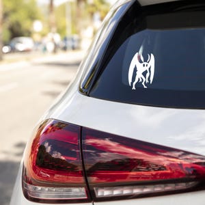 May include: A white vinyl decal of a Mothman creature is affixed to the rear window of a white car. The decal features a stylized silhouette of the cryptid with large wings and a humanoid form. The car's taillight is red.