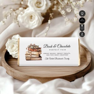 Puede incluir: Un envoltorio de chocolate blanco con una ilustración de acuarela de una pila de libros atados con una cinta. El texto en el envoltorio dice "Book & Chocolate Perfect Pair Sweet Reads. Sweet Treats. See You Next Chapter! The Velvet Bookmark Society."