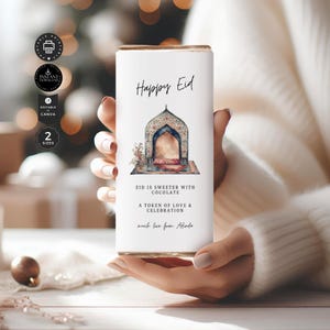 Op de afbeelding: Een witte rechthoekige geschenkdoos met een gouden bovenkant wordt door een persoon vastgehouden. De doos bevat de tekst "Happy Eid" en een illustratie van een boog. Extra tekst luidt "Eid is sweeter with chocolate" en "A token of love & celebration."