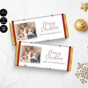 Puede incluir: Dos envoltorios de barra de chocolate con una foto familiar y el texto "Merry Christmas" en escritura roja. Los envoltorios tienen un fondo blanco con detalles en rojo y bordes dorados. También se incluye el texto "FROM THE ALEXANDERS FAMILY".
