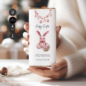 May include: A printable chocolate bar wrapper featuring a pink bunny holding a basket of pink Easter eggs. The wrapper has a pink and white floral garland with pink Easter eggs hanging from it. The text on the wrapper reads "Happy Easter" and "This chocolate is ear-resistible".