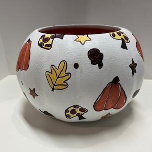 Może przedstawiać: Biała ceramiczna miska o okrągłym kształcie, ozdobiona ręcznie malowanymi ilustracjami o tematyce jesiennej. Projekt obejmuje pomarańczowe dynie, żółte liście, brązowe grzyby i gwiazdy. Wnętrze miski jest czerwonobrązowe.