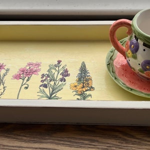 May include: A yellow wooden tray with a white border, featuring a floral design and a small teacup and saucer with a floral pattern.