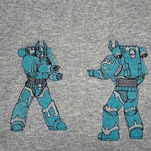 May include: Embroidered design of two blue armored figures on a gray background. The figures are in a fighting stance, with detailed armor and helmets. The design is likely for a t-shirt or other apparel.