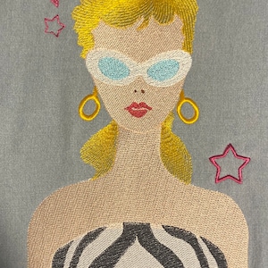 May include: Embroidered portrait of a woman with blonde hair, sunglasses, and gold hoop earrings. The design includes pink stars and a zebra-striped top. The artwork is on a light blue background.
