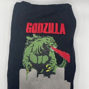 Puede incluir: Prenda negra con un diseño bordado de Godzilla. El diseño incluye la palabra "GODZILLA" en rojo, una figura verde de Godzilla que respira fuego rojo y un paisaje urbano gris. La prenda está doblada.