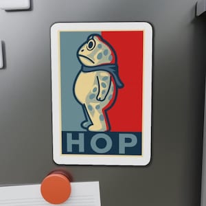 May include: A rectangular magnet featuring a cartoon frog with a blue scarf, set against a red and blue background. The word "HOP" is printed in bold, blue letters at the bottom. The frog is beige with blue spots.
