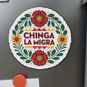 May include: A round white magnet featuring a floral design and the text "CHINGA LA MIGRA" in maroon. The design includes red and yellow flowers, green leaves, and a yellow and red geometric pattern.