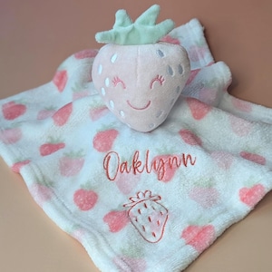 May include: A plush strawberry-shaped baby security blanket. The pink strawberry has a smiling face and green leaves. The white blanket is patterned with pink strawberries and has the name "Oaklynn" embroidered on it.