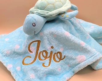 May include: A light blue plush security blanket featuring a soft turtle head and shell attached to the top. The fabric is patterned with small white polka dots and the name JoJo is embroidered in gold script thread on the front surface.